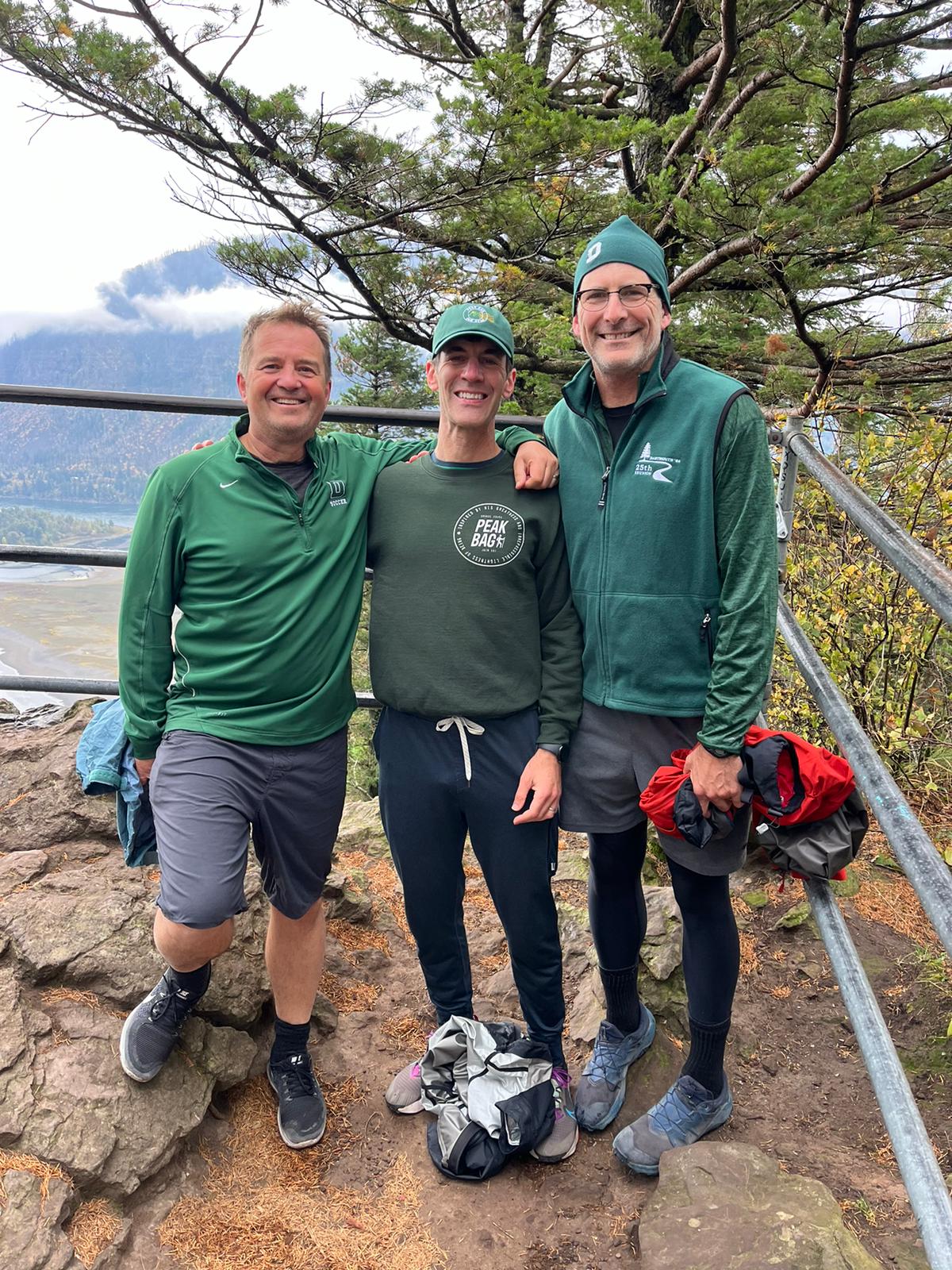 Three people in green and gray outdoor attire standing on a rocky ledge with trees and mountains in the background.
